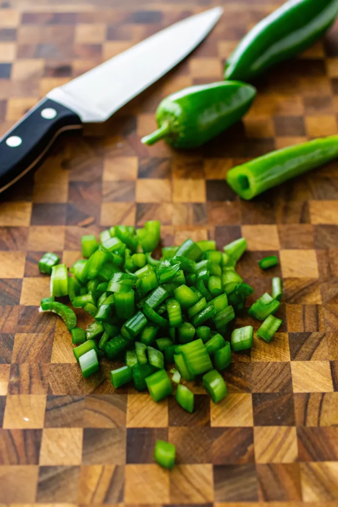 Diced jalapeño peppers on a wooden cutting board with whole jalapeños and a chef's knife