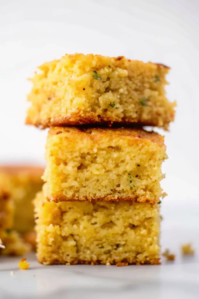Three squares of jalapeño cornbread stacked on top of each other showing moist texture and flecks of green herbs