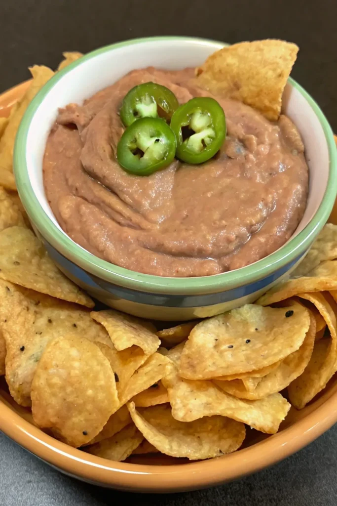 A bowl of bean dip topped with jalapeño slices and a tortilla chip, served in a bowl surrounded by tortilla chips.