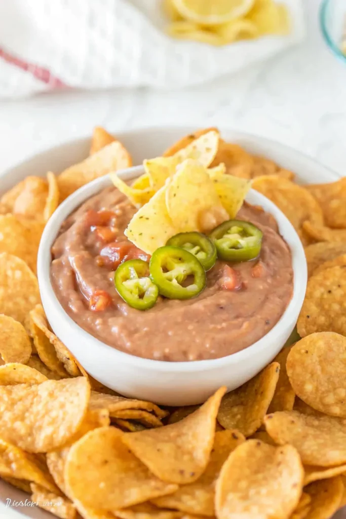 A white bowl of creamy bean dip topped with sliced jalapeños and tortilla chips, surrounded by more tortilla chips on a white plate.