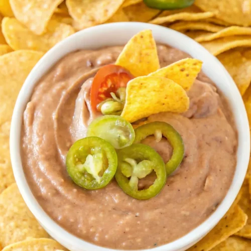 Bean dip in a white bowl topped with jalapeño slices and tortilla chips, surrounded by tortilla chips.